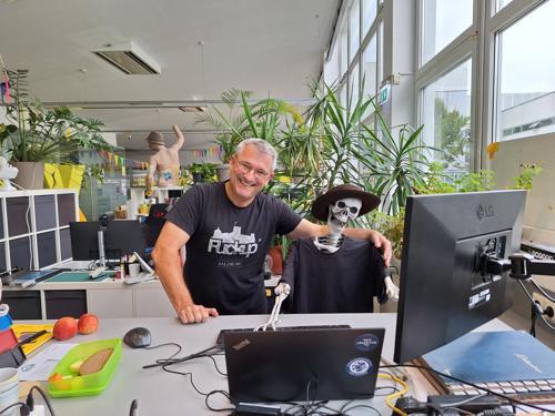 Zu sehen sind 2 lachende Männer an einem IT-Arbeitsplatz. Einer ist jedoch ein Gerippe. Dies ist ein humorvolles Foto als Beispiel für ein Teamfoto in einer Stellenausschreibung