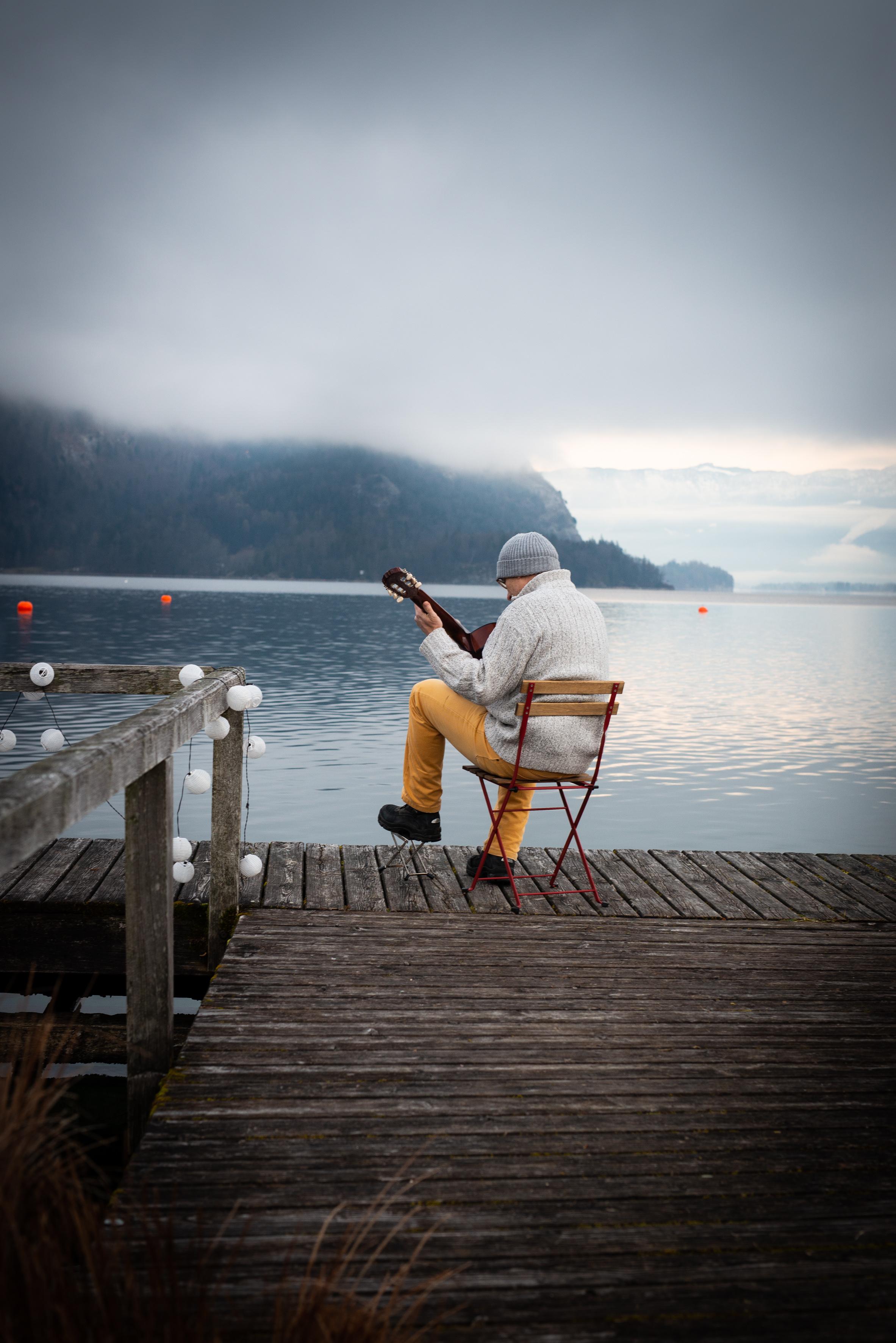 Zu sehen ist ein Gitarrenspieler auf einem Steg an einem herbstlich trüben Alpensee