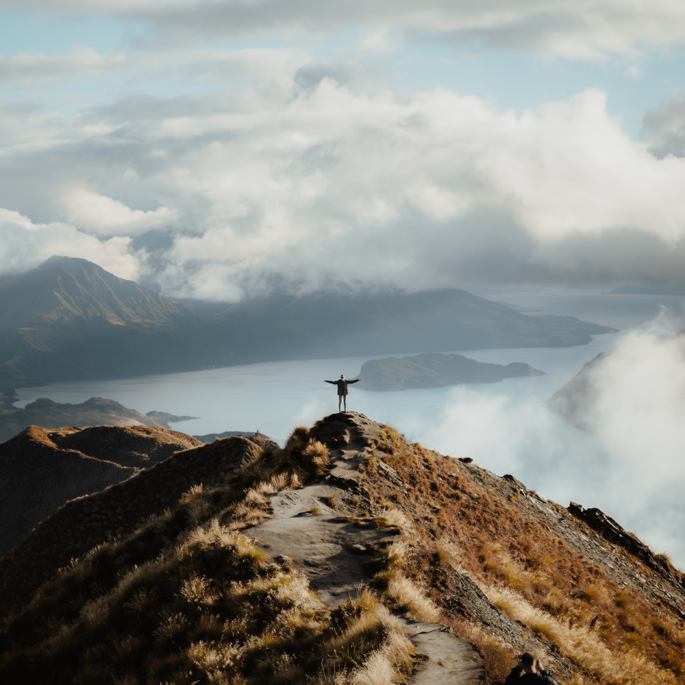 Person, die auf einem Berggipfel steht und die Arme ausstreckt - davor ein atemberaubendes Panorama